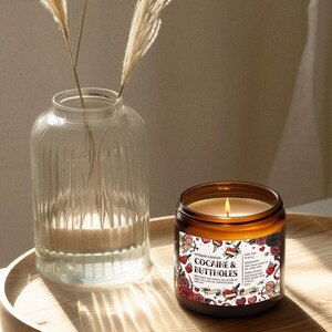 Puede incluir: Una vela encendida en un tarro de cristal marr&oacute;n con el texto "COCAINE & BUTTHOLES" en una etiqueta con rosas e ilustraciones de cupido. La vela est&aacute; sobre una bandeja de madera junto a un jarr&oacute;n de cristal transparente con plantas secas.