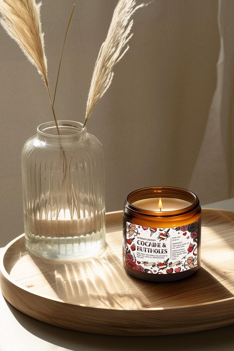Puede incluir: Una vela encendida en un tarro de cristal marr&oacute;n con el texto "COCAINE & BUTTHOLES" en una etiqueta con rosas e ilustraciones de cupido. La vela est&aacute; sobre una bandeja de madera junto a un jarr&oacute;n de cristal transparente con plantas secas.