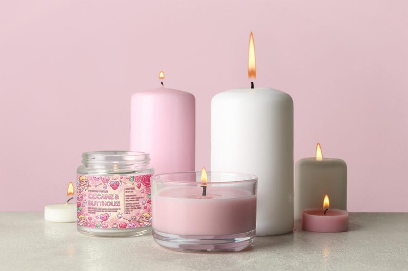 May include: A collection of lit candles in various sizes and shapes, including a jar candle with the text "COCAINE & BUTTHOLES". The candles are in shades of pink, white, and gray, with flames. The background is a solid pink.