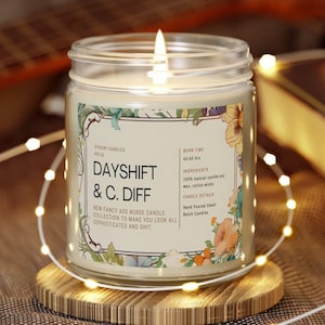 May include: A burning candle in a glass jar, labelled "DAYSHIFT & C. DIFF", with floral designs. The label states it's a soy wax candle with a 45-50 hour burn time. It rests on a wooden coaster, encircled by decorative fairy lights.