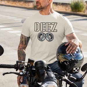 May include: A light beige t-shirt with the word "DEEZ" in a black outlined font and a graphic of two nuts. The person is wearing blue jeans and is sitting on a black motorcycle. A black helmet is held in the person's hand.