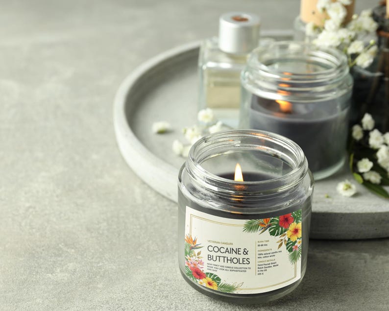 May include: A lit, dark gray candle in a clear glass jar with a label that reads "Cocaine & Buttholes." The label features tropical flowers and foliage. Other candles and a perfume bottle are in the background on a gray tray.