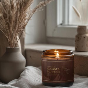 May include: A lit candle in a brown glass jar with a label that reads "COCAINE & BUTTHOLES". The candle sits on a white cloth, with a vase of dried plants to the left and a window in the background. The candle's flame glows brightly.