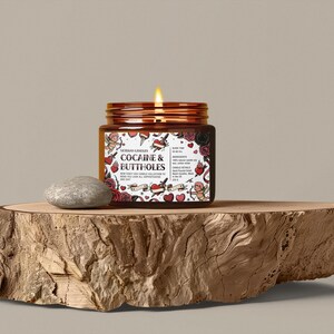 Puede incluir: Una vela encendida en un frasco marr&oacute;n con tapa de cobre, etiquetada "Cocaine & Buttholes" con gr&aacute;ficos estilo tatuaje. La vela est&aacute; sobre una losa de madera con una piedra gris al lado.