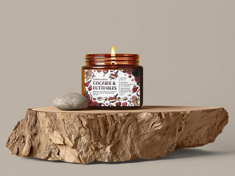 Puede incluir: Una vela encendida en un frasco marr&oacute;n con tapa de cobre, etiquetada "Cocaine & Buttholes" con gr&aacute;ficos estilo tatuaje. La vela est&aacute; sobre una losa de madera con una piedra gris al lado.