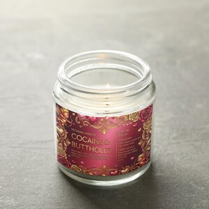 Peut inclure: Une bougie en pot de verre transparent avec une m&egrave;che allum&eacute;e. L'&eacute;tiquette porte le texte "COCAINE & BUTTHOLES" dans une police d&eacute;corative, entour&eacute;e d'un motif floral rouge et or. La bougie est sur une surface grise.