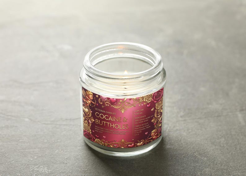 Peut inclure: Une bougie en pot de verre transparent avec une m&egrave;che allum&eacute;e. L'&eacute;tiquette porte le texte "COCAINE & BUTTHOLES" dans une police d&eacute;corative, entour&eacute;e d'un motif floral rouge et or. La bougie est sur une surface grise.