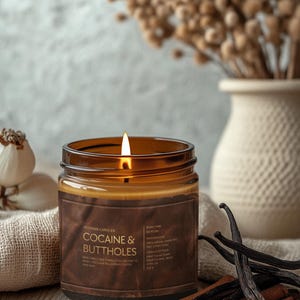 May include: A lit brown glass jar candle with a label that reads "COCAINE & BUTTHOLES". The candle is part of the "Veteran Candles" collection. The candle is surrounded by cinnamon sticks, vanilla beans, and a vase with dried flowers.