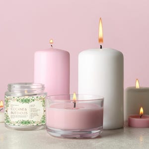 Op de afbeelding: Een verzameling brandende kaarsen in verschillende maten en vormen, waaronder een potkaars met de tekst "COCAINE & BUTTHOLES". De kaarsen zijn in roze, witte en grijze tinten, met vlammen tegen een roze achtergrond.