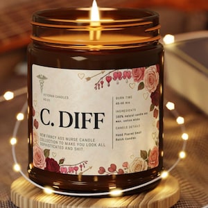 May include: A lit brown glass jar candle with a label featuring floral designs and the text "C. DIFF." The label also includes the text "New Fancy Ass Nurse Candle Collection" and "Veteran Candles." The candle sits on a wooden coaster.