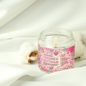 May include: A lit, clear glass jar candle with a pink label featuring cartoon cherubs, hearts, and roses. The text on the label reads "COCAINE & BUTTHOLES". The candle sits on a white fabric with cotton buds.