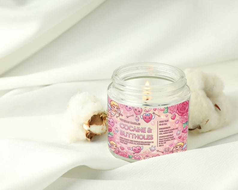 May include: A lit, clear glass jar candle with a pink label featuring cartoon cherubs, hearts, and roses. The text on the label reads "COCAINE & BUTTHOLES". The candle sits on a white fabric with cotton buds.