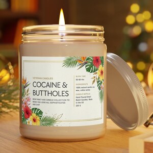 May include: A lit candle in a glass jar with a lid. The label reads "COCAINE & BUTTHOLES" with floral accents. The candle is part of the "NEW FANCY ASS CANDLE COLLECTION". The jar contains 225 grams of wax.