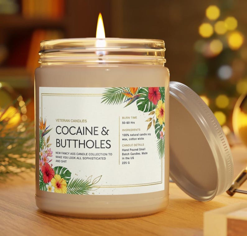 May include: A lit candle in a glass jar with a lid. The label reads "COCAINE & BUTTHOLES" with floral accents. The candle is part of the "NEW FANCY ASS CANDLE COLLECTION". The jar contains 225 grams of wax.