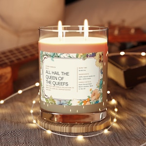 Puede incluir: Un tarro de vidrio para velas con una etiqueta de diseño floral que dice "All Hail the Queen of the Queefs". La etiqueta también incluye detalles sobre la vela, como el tiempo de combustión, los ingredientes y los detalles de la vela. La vela está encendida y tiene dos mechas.