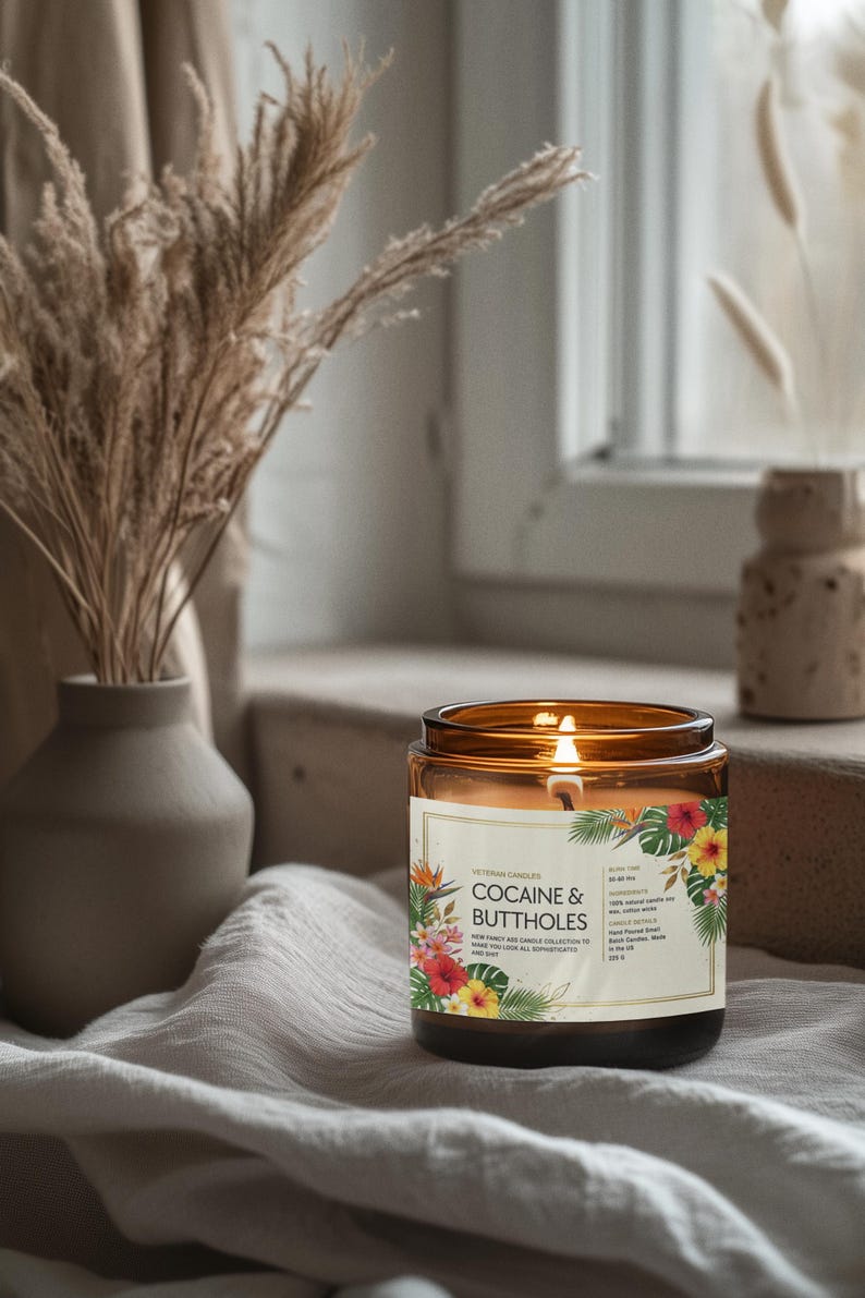May include: A lit candle in a brown glass jar with a label that reads "Cocaine & Buttholes" surrounded by tropical flowers. The candle sits on a white cloth near a window, with a vase of dried plants in the background.