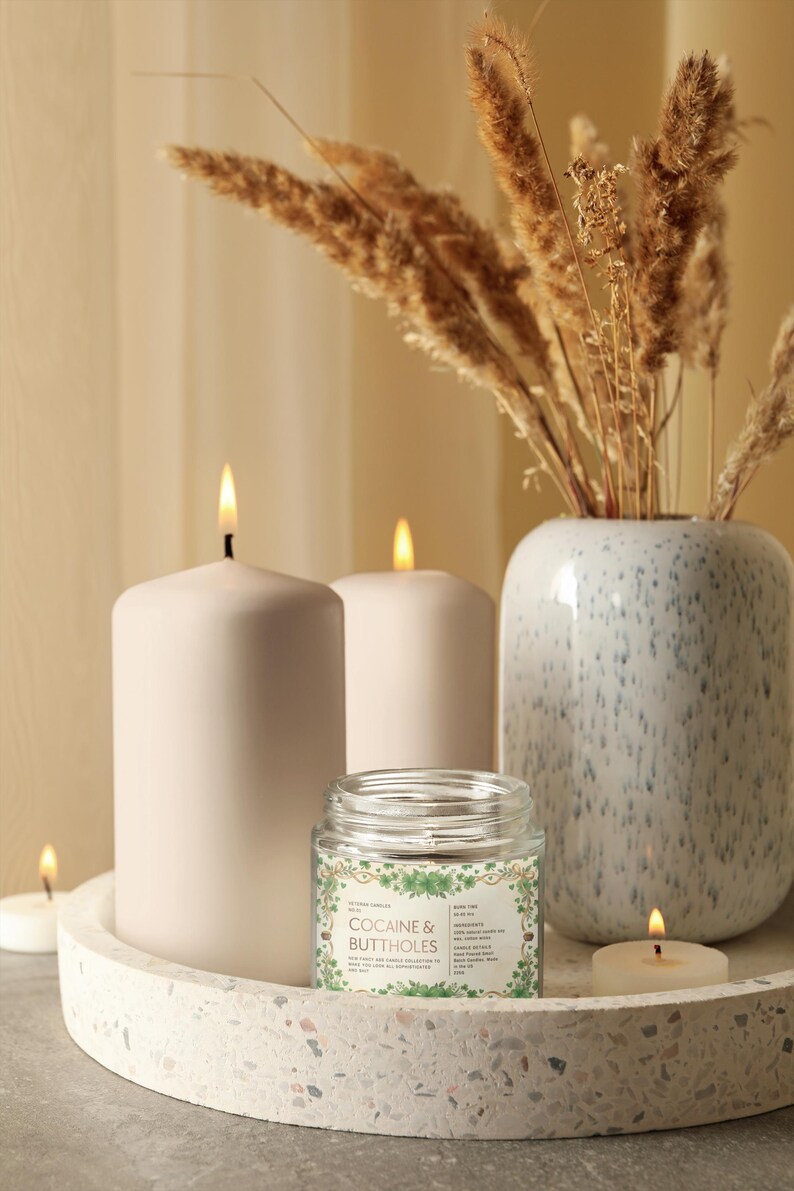 Op de afbeelding: Een decoratieve opstelling met brandende kaarsen en een kleine potkaars met de tekst "COCAINE & BUTTHOLES". De kaarsen staan op een terrazzo dienblad, met een vaas met gedroogde planten op de achtergrond. De sc&egrave;ne heeft een warme sfeer.