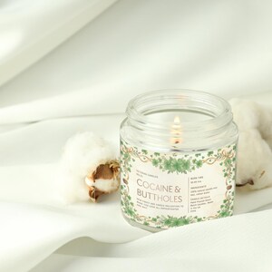 Op de afbeelding: Een brandende, heldere glazen potkaars met een label met de tekst "COCAINE & BUTTHOLES" omgeven door groene bloemmotieven. De kaars staat op een witte doek naast katoenbollen. De kaars maakt deel uit van een nieuwe, chique kaarsencollectie.