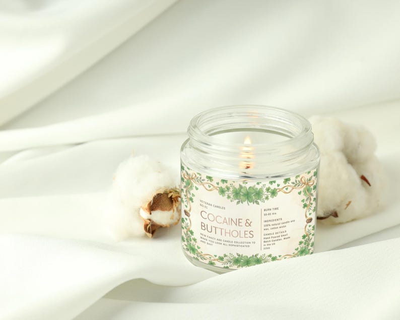 Op de afbeelding: Een brandende, heldere glazen potkaars met een label met de tekst "COCAINE & BUTTHOLES" omgeven door groene bloemmotieven. De kaars staat op een witte doek naast katoenbollen. De kaars maakt deel uit van een nieuwe, chique kaarsencollectie.