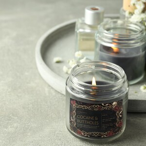 May include: A lit, dark gray candle in a clear glass jar with a label that reads "COCAINE & BUTTHOLES". The label has a decorative border with skulls and roses. Other candles and a perfume bottle are in the background.