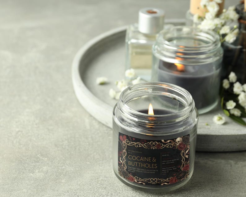 May include: A lit, dark gray candle in a clear glass jar with a label that reads "COCAINE & BUTTHOLES". The label has a decorative border with skulls and roses. Other candles and a perfume bottle are in the background.