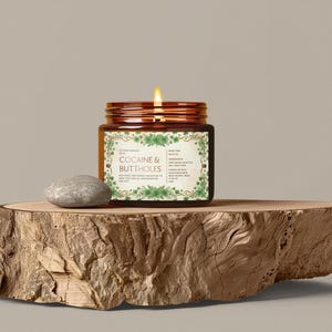 Może przedstawiać: Zapachowa świeca w szklanym, brązowym słoiku z napisem "COCAINE & BUTTHOLES" na drewnianej desce. Obok świecy leży mały szary kamień. Świeca ma brązową pokrywę i etykietę z zielonymi kwiatowymi akcentami.