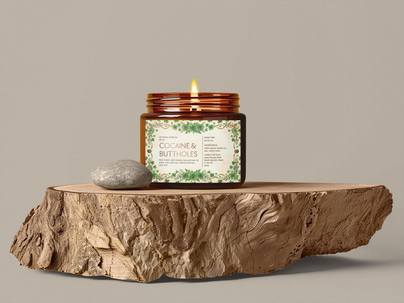 Może przedstawiać: Zapachowa świeca w szklanym, brązowym słoiku z napisem "COCAINE & BUTTHOLES" na drewnianej desce. Obok świecy leży mały szary kamień. Świeca ma brązową pokrywę i etykietę z zielonymi kwiatowymi akcentami.