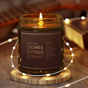 May include: A lit candle in a glass jar with a brown label that reads "COCAINE & BUTTHOLES." The candle sits on a wooden coaster, surrounded by string lights. The jar is filled with wax and has a burn time of 50-60 hrs.