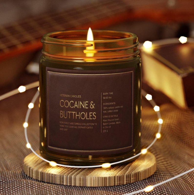 May include: A lit candle in a glass jar with a brown label that reads "COCAINE & BUTTHOLES." The candle sits on a wooden coaster, surrounded by string lights. The jar is filled with wax and has a burn time of 50-60 hrs.