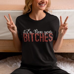 May include: Black t-shirt with the phrase "it's a New year BITCHES" in white and red glitter lettering. The shirt is a crew neck style.