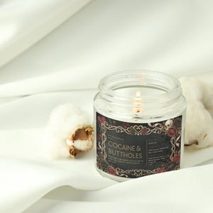 Pu&ograve; includere: Una candela accesa in un barattolo di vetro trasparente con un'etichetta nera che recita "COCAINE & BUTTHOLES" in testo bianco. L'etichetta ha un bordo decorativo con rose e teschi. La candela &egrave; su una superficie bianca con batuffoli di cotone.