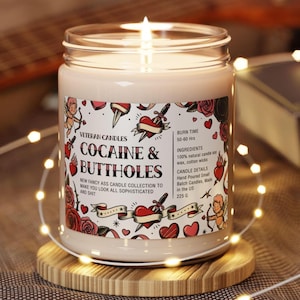 May include: A lit candle in a glass jar with a label that reads "COCAINE & BUTTHOLES." The label features tattoo-style graphics of hearts, roses, and cherubs. The candle is on a wooden coaster, surrounded by string lights.