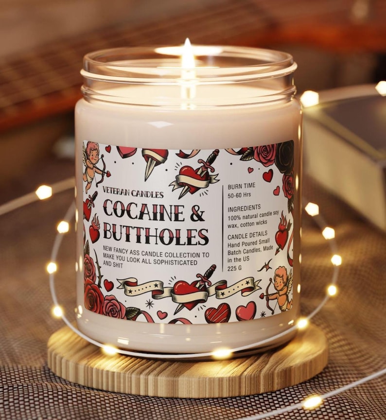 May include: A lit candle in a glass jar with a label that reads "COCAINE & BUTTHOLES." The label features tattoo-style graphics of hearts, roses, and cherubs. The candle is on a wooden coaster, surrounded by string lights.