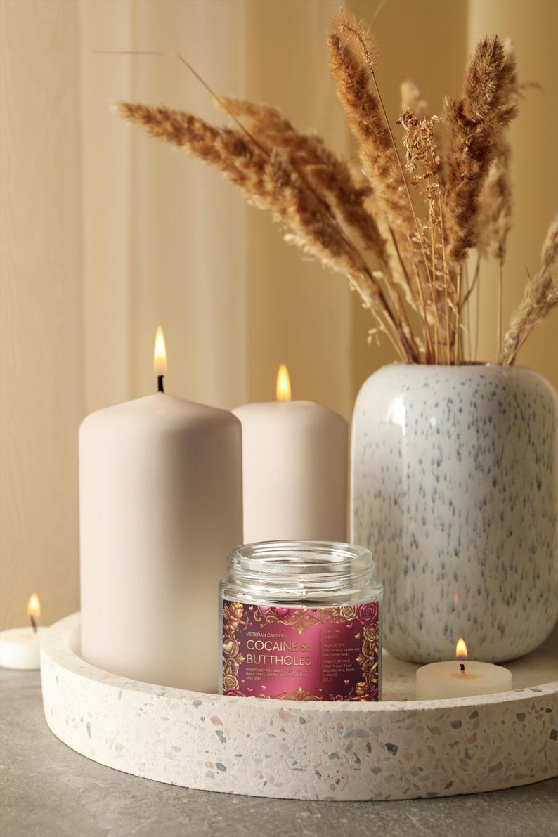 Peut inclure: Composition d&eacute;corative avec trois bougies blanches allum&eacute;es, une petite bougie en pot avec une &eacute;tiquette et un vase avec des plantes s&eacute;ch&eacute;es. L'&eacute;tiquette de la bougie en pot indique "COCAINE & BUTTHOLES". Les objets sont sur un plateau d&eacute;coratif.