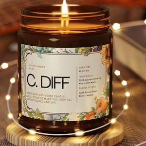 May include: A burning brown glass jar candle with a floral label. The label says "C. DIFF" and "NEW FANCY ASS NURSE CANDLE COLLECTION TO MAKE YOU LOOK ALL SOPHISTICATED AND SHIT." The candle is on a wooden coaster, with fairy lights around it.