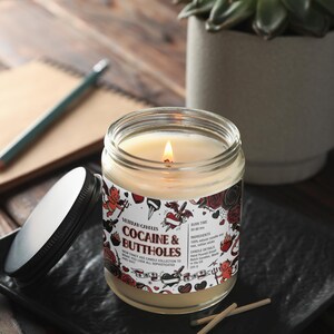 Może przedstawiać: Zapalone świeca w szklanym słoiku z czarną pokrywką, z napisem "Cocaine & Buttholes". Etykieta zawiera ilustracje i tekst w stylu tatuażu. Świeca stoi na czarnej kwadratowej tacy z zapałkami w pobliżu.