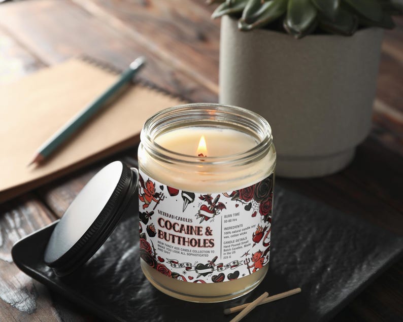 Może przedstawiać: Zapalone świeca w szklanym słoiku z czarną pokrywką, z napisem "Cocaine & Buttholes". Etykieta zawiera ilustracje i tekst w stylu tatuażu. Świeca stoi na czarnej kwadratowej tacy z zapałkami w pobliżu.