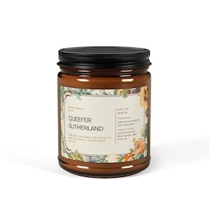 May include: A brown glass candle jar with a black lid. The label reads "Sour Candles" and "Queefer Sutherland". The candle is decorated with a floral design and has a burn time of 30-80 hours.