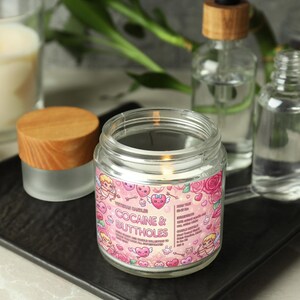 May include: A lit glass jar candle with a pink label that reads "Cocaine & Buttholes". The label features cartoon cherubs, hearts, and roses. Other items include a small jar with a wooden lid, and glass bottles.