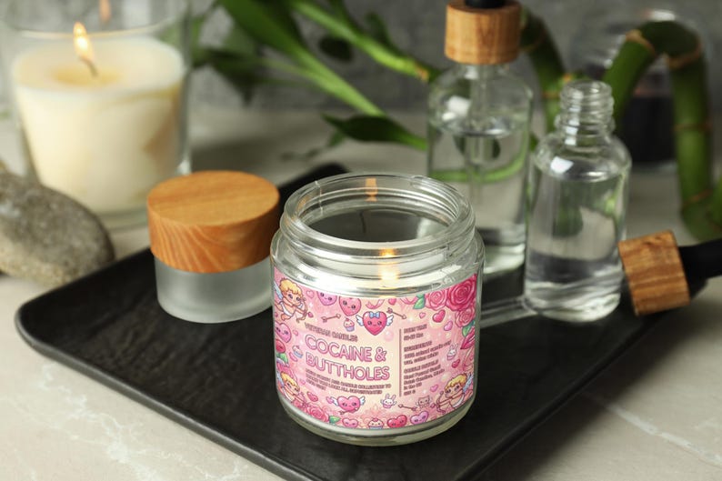 May include: A lit glass jar candle with a pink label that reads "Cocaine & Buttholes". The label features cartoon cherubs, hearts, and roses. Other items include a small jar with a wooden lid, and glass bottles.
