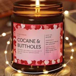 May include: A lit candle in a brown glass jar with a pink label featuring the text "COCAINE & BUTTHOLES". The label is decorated with red and white hearts. The candle sits on a wooden coaster, surrounded by string lights.