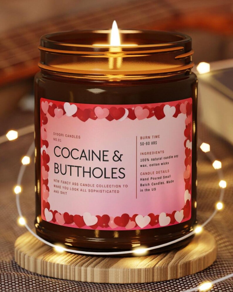 May include: A lit candle in a brown glass jar with a pink label featuring the text "COCAINE & BUTTHOLES". The label is decorated with red and white hearts. The candle sits on a wooden coaster, surrounded by string lights.