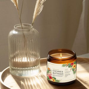 May include: A lit candle in a brown glass jar with a label that reads "COCAINE & BUTTHOLES" with floral accents. A clear glass vase with pampas grass sits to the left on a wooden tray.