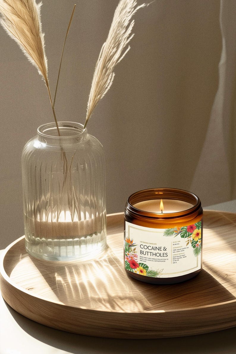 May include: A lit candle in a brown glass jar with a label that reads "COCAINE & BUTTHOLES" with floral accents. A clear glass vase with pampas grass sits to the left on a wooden tray.