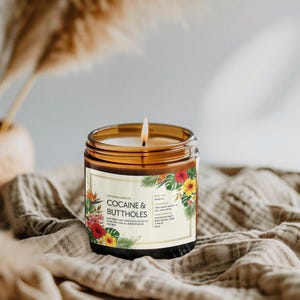Może przedstawiać: Zapalone świeca w brązowym szklanym słoiku z etykietą z napisem "COCAINE & BUTTHOLES" otoczoną tropikalnymi kwiatami. Świeca stoi na teksturowanej, beżowej powierzchni. Świeca jest częścią linii "NEW FANCY ASS CANDLE COLLECTION TO MAKE YOU LOOK ALL SOPHISTICATED".