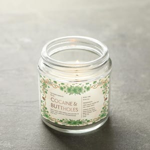 Op de afbeelding: Een brandende, heldere glazen potkaars met een label met de tekst "Cocaine & Buttholes". Het label heeft een groen en goudkleurig bloemmotief. De kaars staat op een grijze ondergrond.