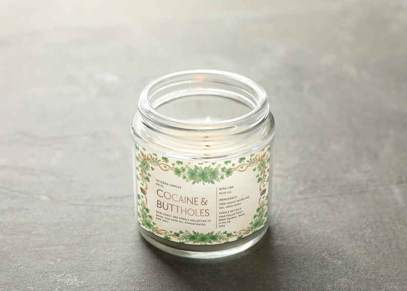 Op de afbeelding: Een brandende, heldere glazen potkaars met een label met de tekst "Cocaine & Buttholes". Het label heeft een groen en goudkleurig bloemmotief. De kaars staat op een grijze ondergrond.