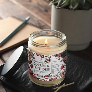 May include: A lit glass jar candle with a label that reads "Cocaine & Buttholes" surrounded by tattoo-style graphics. The candle sits on a black square dish with the lid beside it. A pencil and notebook are in the background.