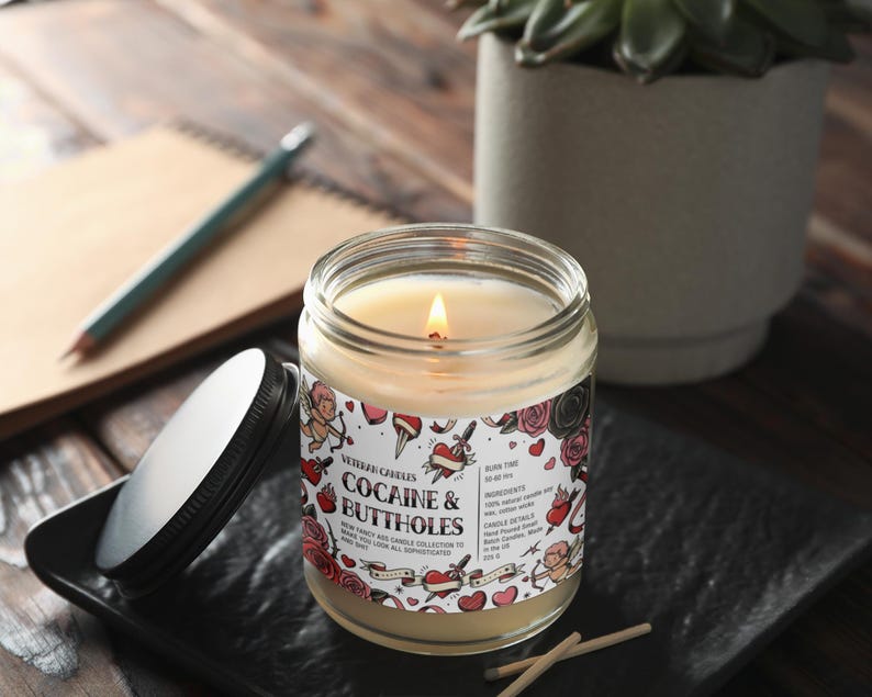May include: A lit glass jar candle with a label that reads "Cocaine & Buttholes" surrounded by tattoo-style graphics. The candle sits on a black square dish with the lid beside it. A pencil and notebook are in the background.