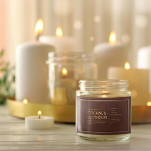 May include: A lit candle in a glass jar with a brown label that reads "COCAINE & BUTTHOLES" is in focus. Several other lit candles of varying sizes are in the background, creating a warm, inviting ambiance. The candles are on a wooden surface.
